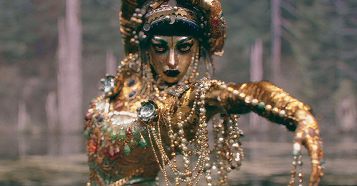 A heavily jeweled woman rises out of a lake and dances